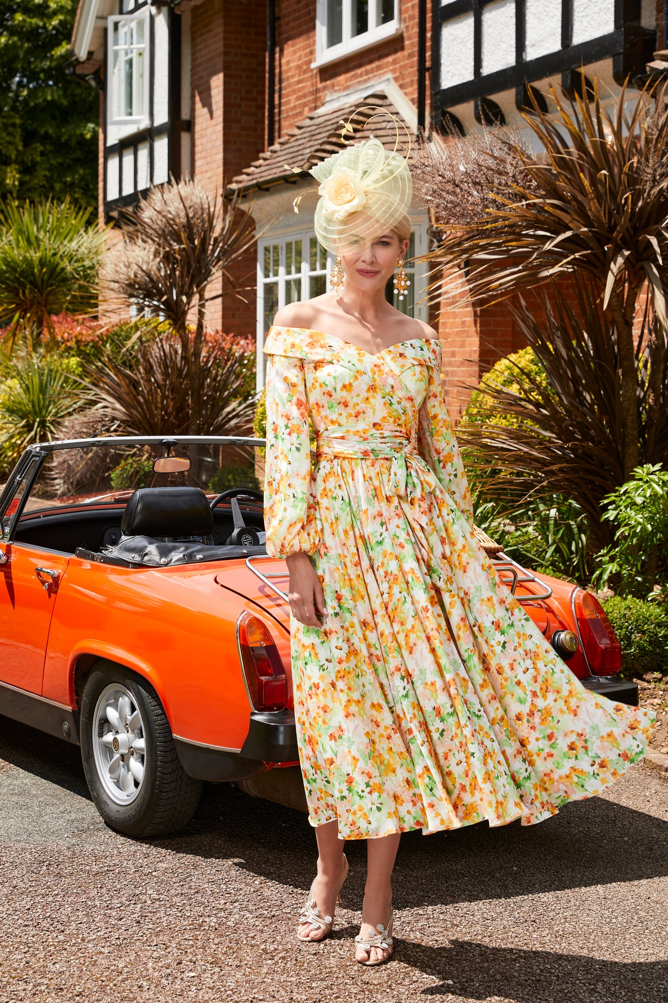 Woman in vivid yellow and orange floral a-line dress with off the shoulder detail and long blouson sleeves standing in front of vintage orange car